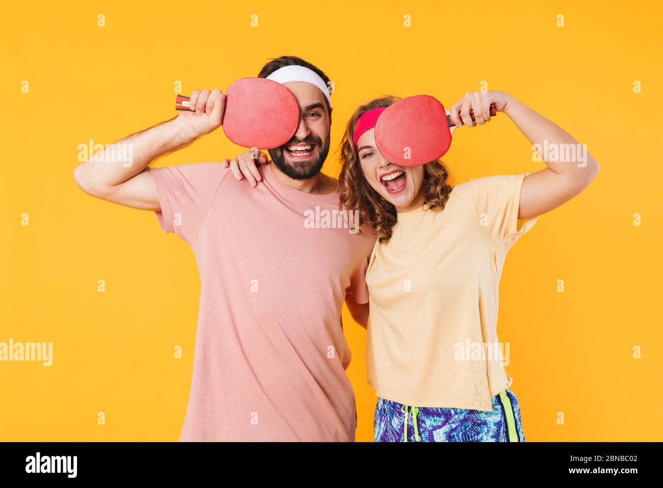 Ritratto di giovane coppia sportiva felice con archetti che sorridono e tengono racchette da ping pong isolate su sfondo giallo Foto Stock