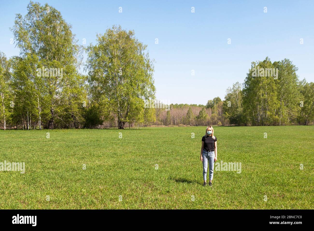 Ragazza bionda in T-shirt nera e jeans blu con una maschera medica sul viso è solo nel campo contro uno sfondo di alberi e erba. Autoisolati Foto Stock