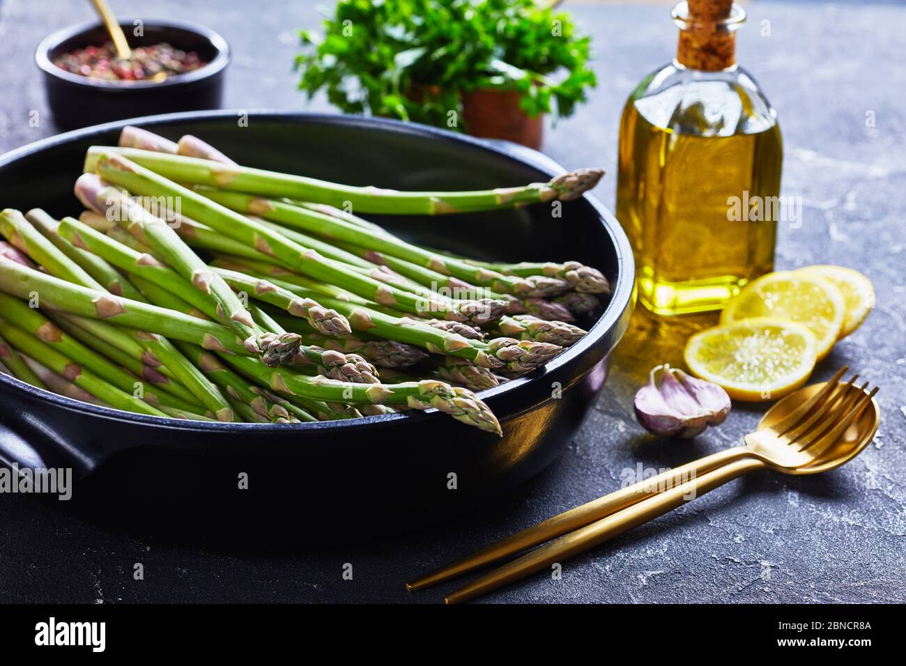 primo piano di asparagi verdi freschi in teglia nera con posate dorate, fette di limone, peppercorne e bottiglia di olio d'oliva sulla linguetta grigia in cemento Foto Stock