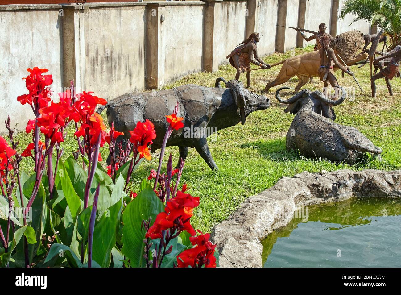 Sculture all'aperto, animali, leoni tribesmen spiearing, fiori rossi, laghetto, acqua, arte, Centro Culturale, Africa, Arusha, Tanzania Foto Stock