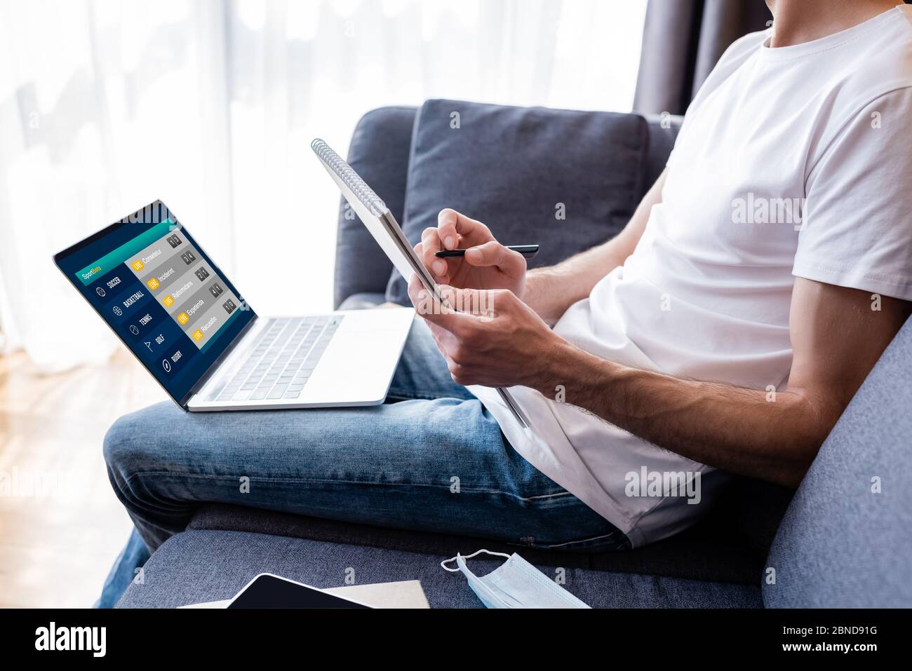 Vista ritagliata della scrittura uomo sul notebook vicino al portatile con scommesse sportive sullo schermo e maschera medica sul divano Foto Stock