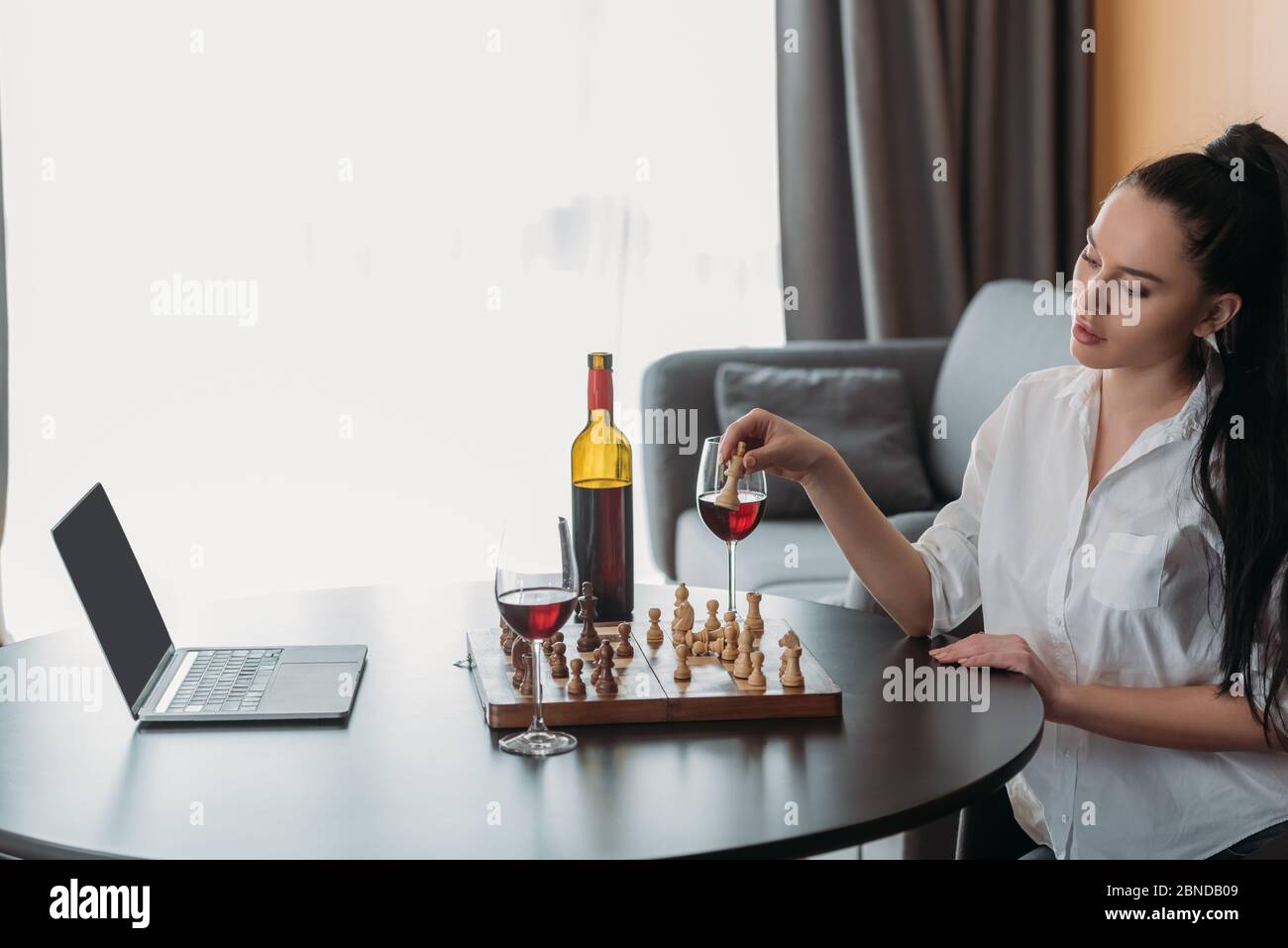 bella donna che gioca a scacchi vicino al computer portatile con schermo vuoto e vino rosso sul tavolo Foto Stock