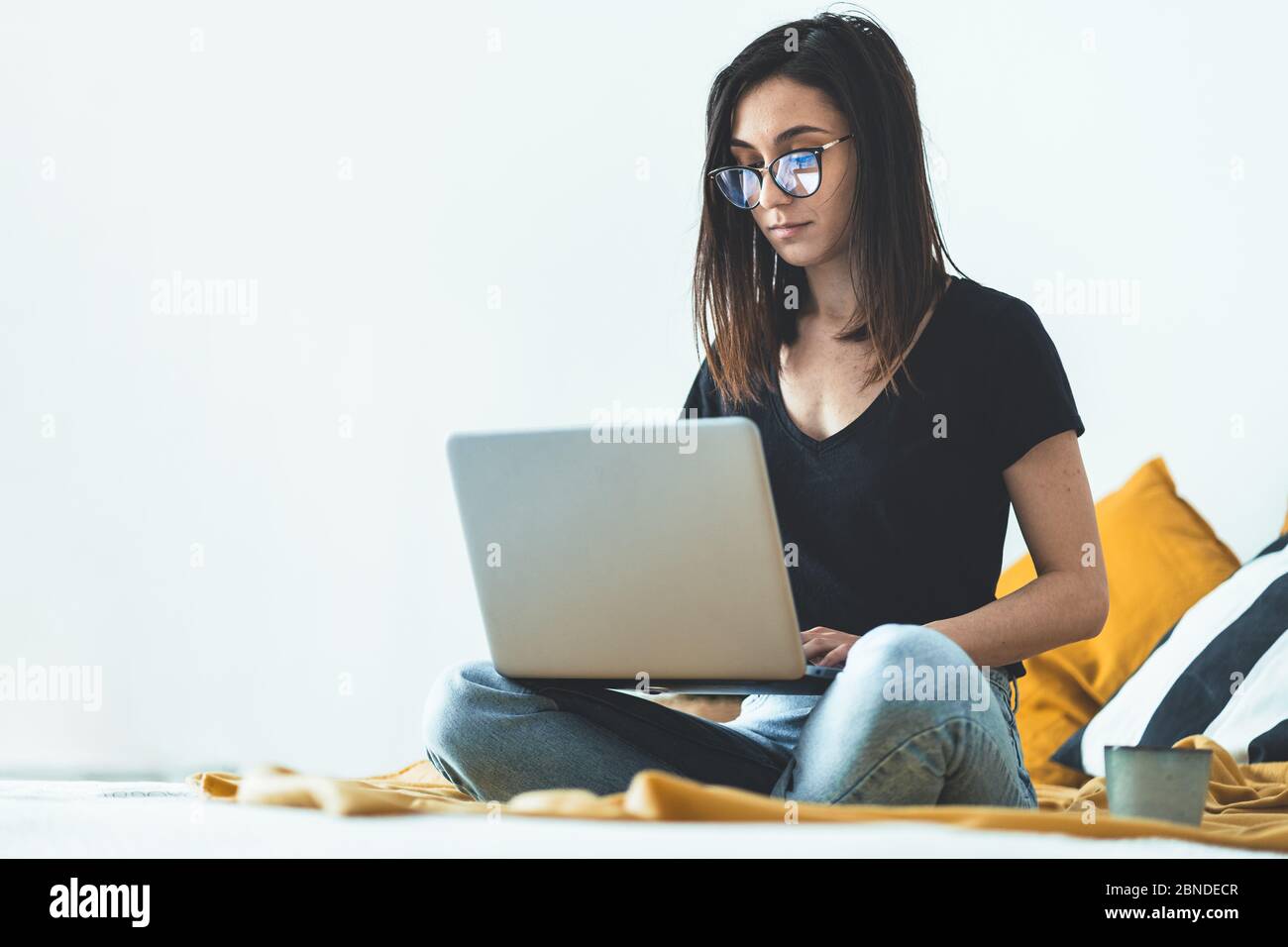 Attraente giovane donna che indossa occhiali da vista che lavorano da home.Female imprenditore seduto sul letto e che lavora dal computer portatile mentre beve caffè Foto Stock