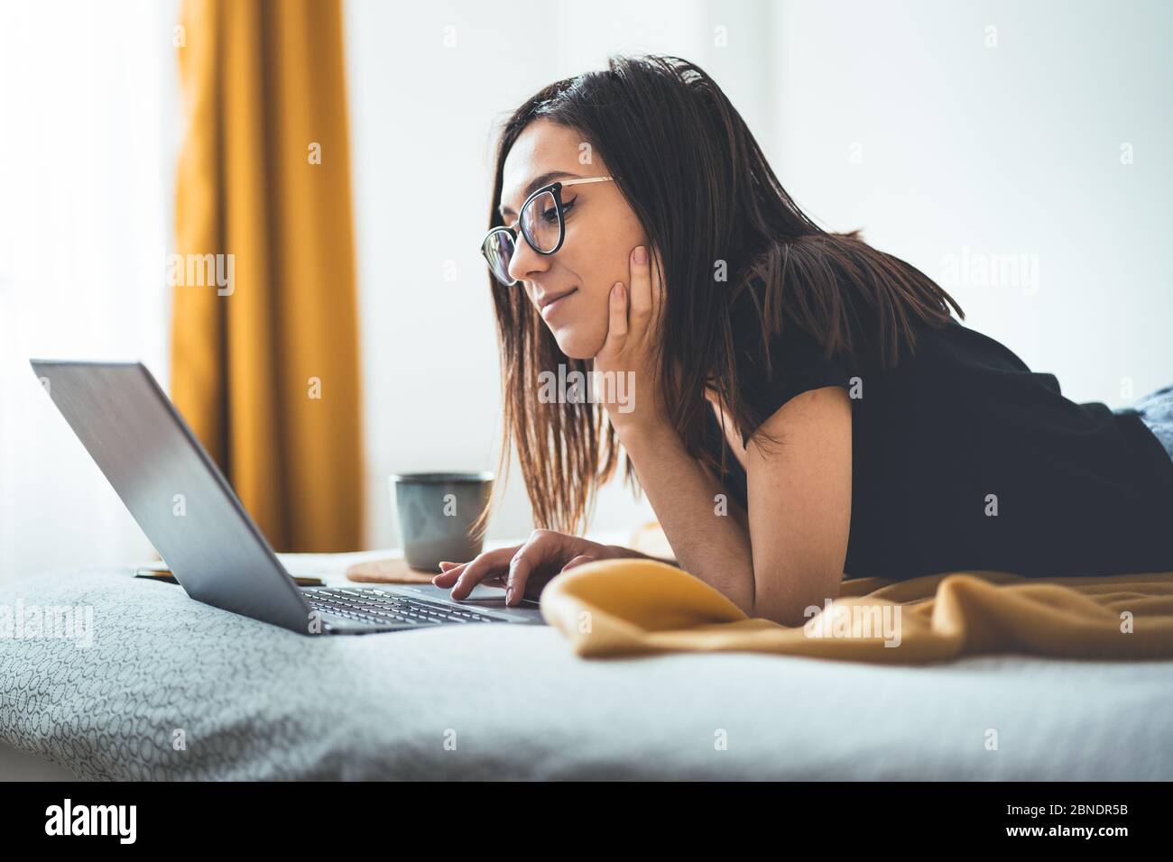 Sorridente bella donna sdraiata sul letto utilizzando il computer portatile che comunica online a casa, felice ragazza che indossa occhiali e digitando sul computer, godendo intern Foto Stock