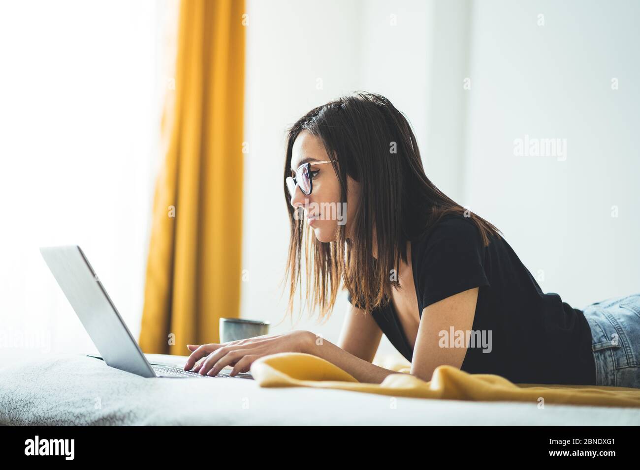 Bella donna che indossa occhiali e lavora su computer portatile. Femmina concentrata che si trova a letto e lavora da casa. Stile di vita e corona freelance Foto Stock