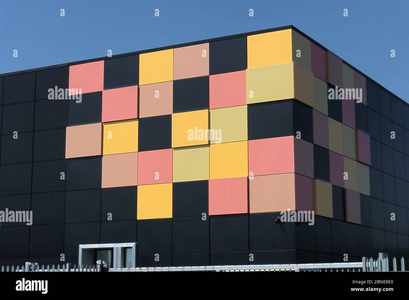 Edificio cubico nero opaco con piastrelle gialle e rosse in un angolo come design grafico dietro le ringhiere di sicurezza in metallo, data center cielo blu intenso Foto Stock