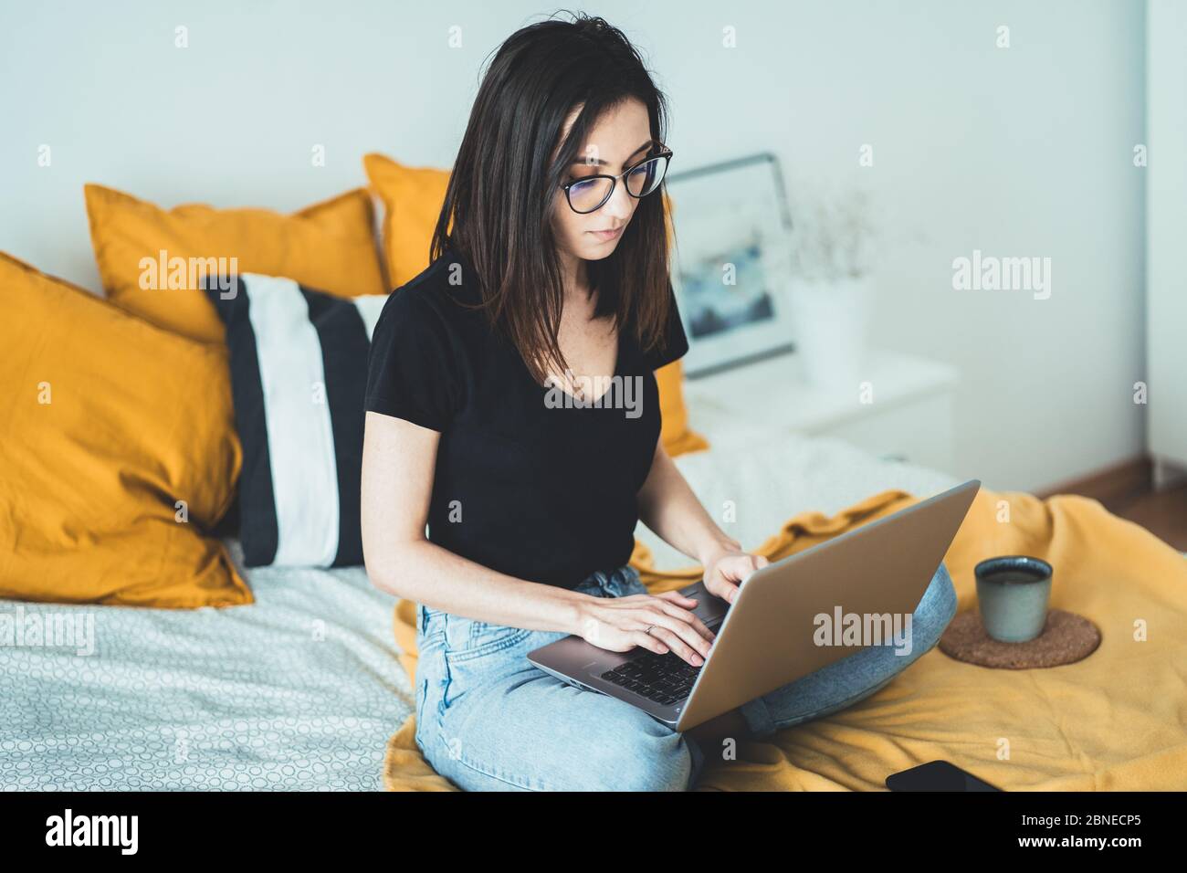 Donna casual che lavora su un computer portatile seduto sul letto nel letto. Occhiali da vista da donna che si rilassano e bevono una tazza di caffè o tè caldo utilizzando la Foto Stock
