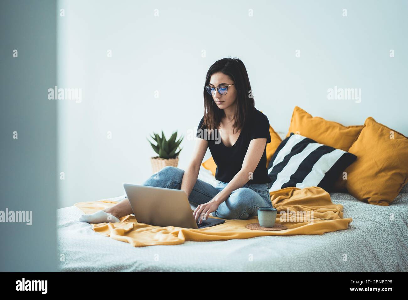 Attraente giovane donna che indossa occhiali da vista che lavora da casa. Donna imprenditore seduto a letto e lavorando da computer portatile mentre beve morni Foto Stock
