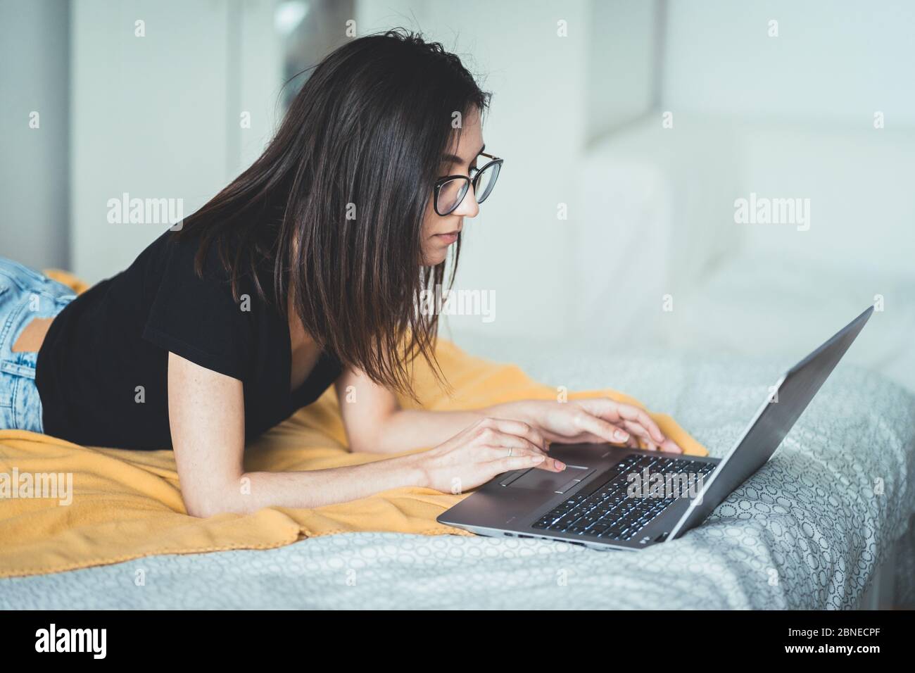 Bella donna brunetta che indossa occhiali e lavora su computer portatile. Donna concentrata che si trova a letto a casa scrivendo e utilizzando la tastiera del notebook Foto Stock