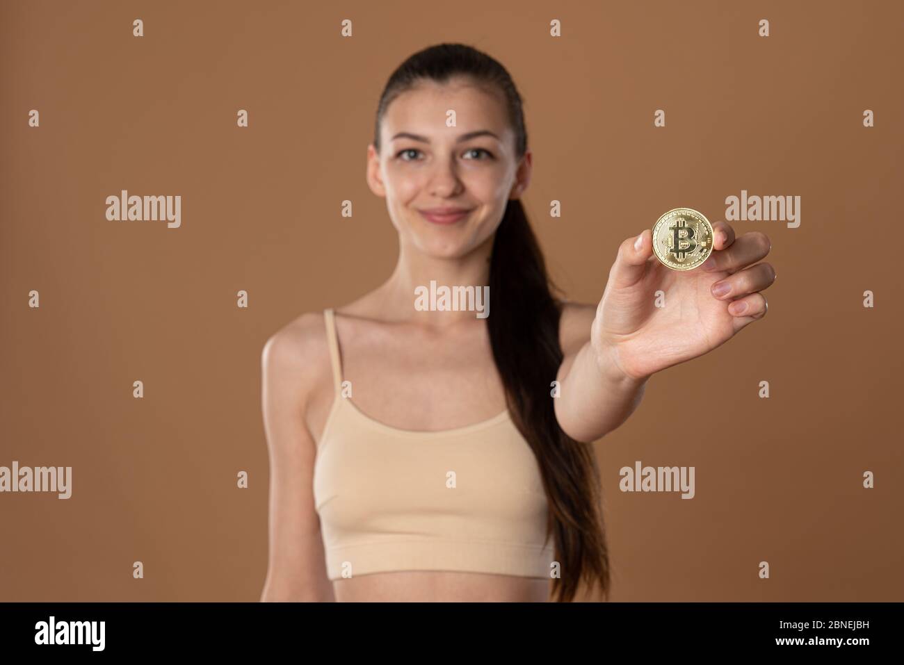 Vita in su foto di bruna ragazza felice in bra beige che tiene moneta d'oro bitcoin e guardando la fotocamera Foto Stock