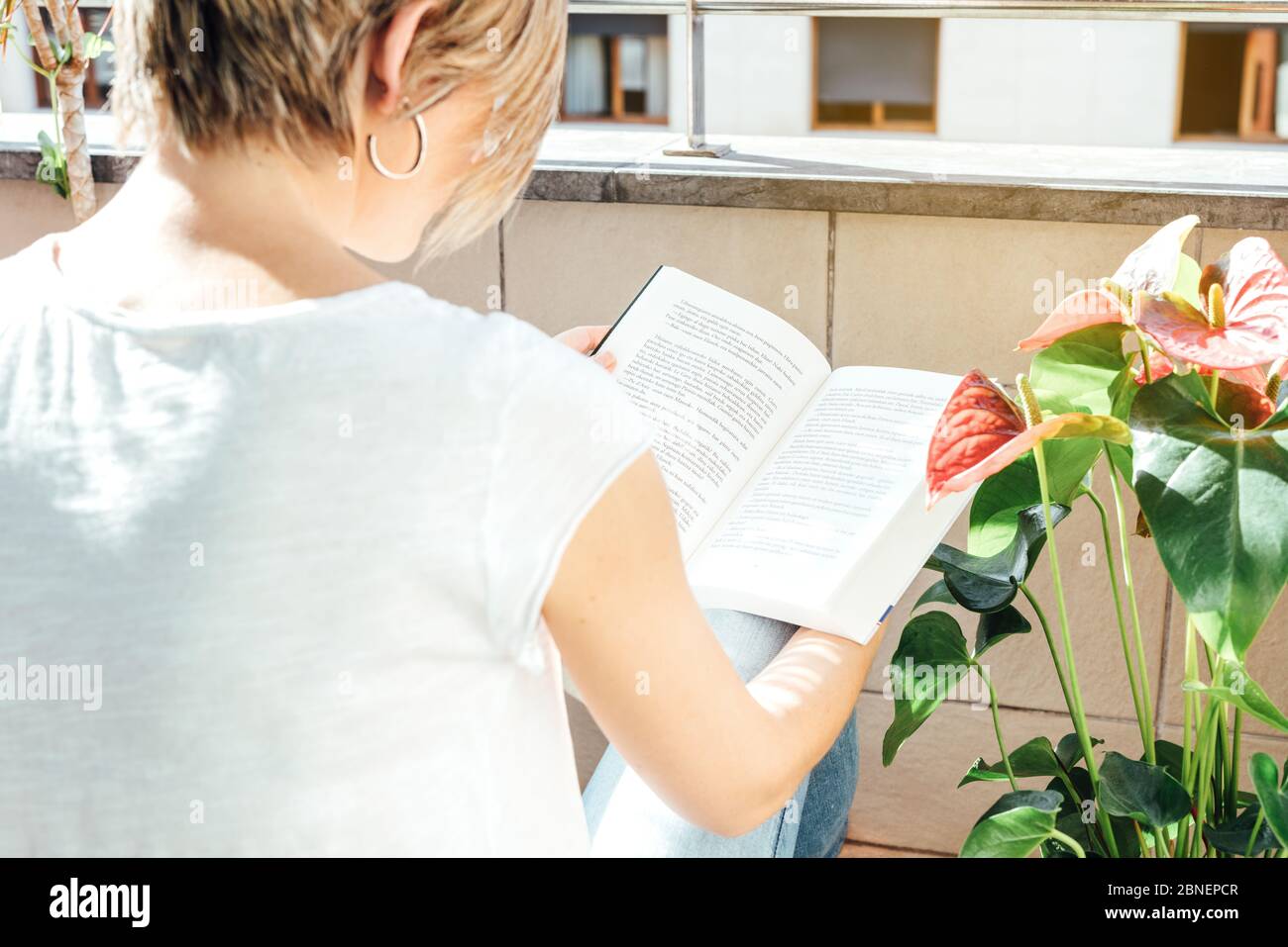 donna bionda che legge un libro seduto sul pavimento del balcone circondato da piante al tramonto e confinato alla sua casa a causa della pandemia del coronavirus Foto Stock