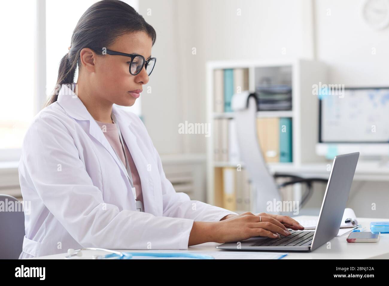 Dottore in occhiali e cappotto bianco seduto al tavolo e concentrandosi sul suo lavoro su computer portatile Foto Stock