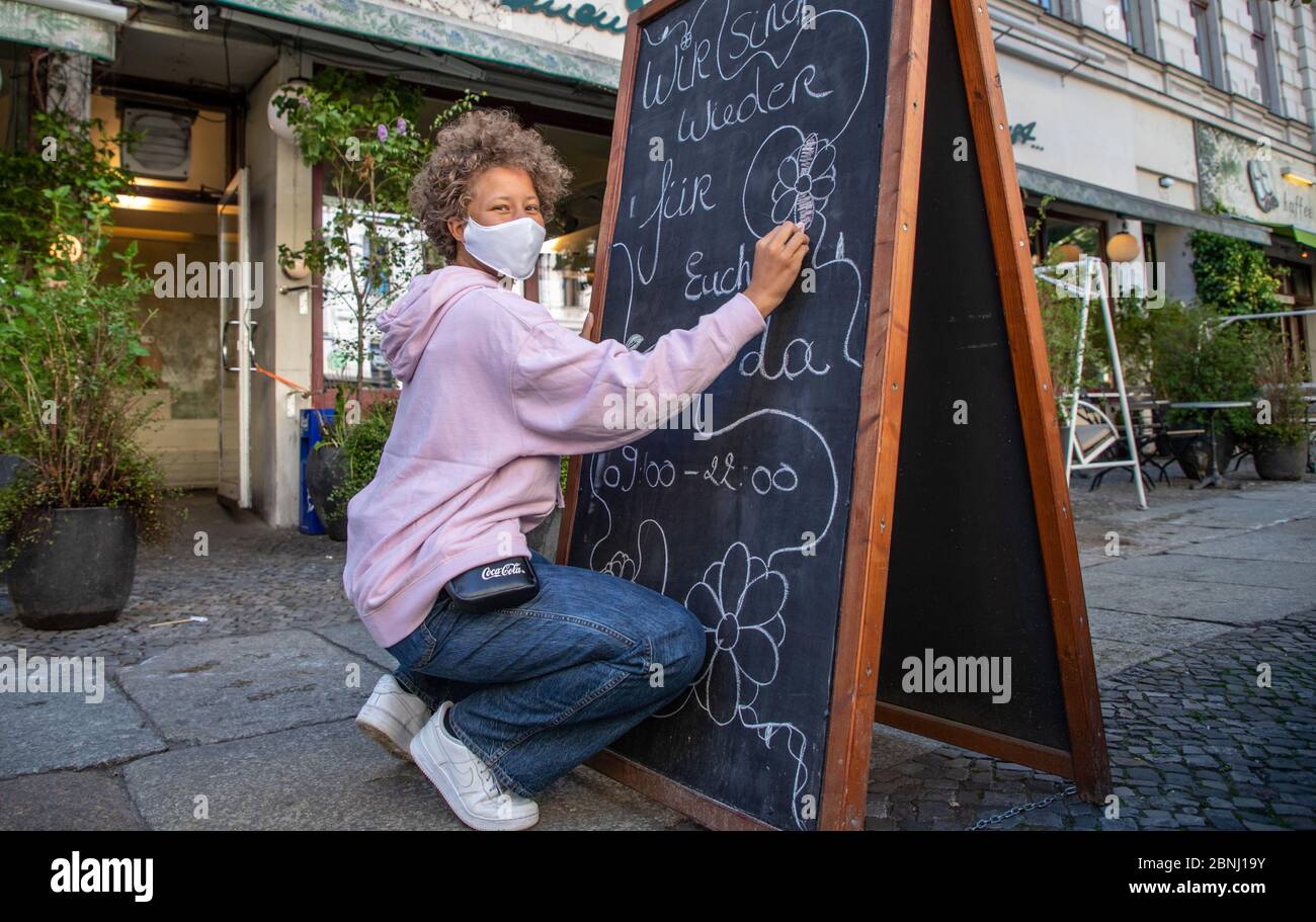 Berlino, Germania. 15 maggio 2020. Il membro del personale Kiki del caffè 'una domenica di agosto' indossa una guardia quando scrive 'Siamo qui per voi di nuovo' su una lavagna davanti al ristorante. Dal 15.05., ristoranti e caffè a Berlino possono riaprire in determinate condizioni. Gli alimenti preparati di fresco possono essere consumati anche in loco, a condizione che venga osservata la distanza minima. Credit: Andreas Gora/dpa/Alamy Live News Foto Stock