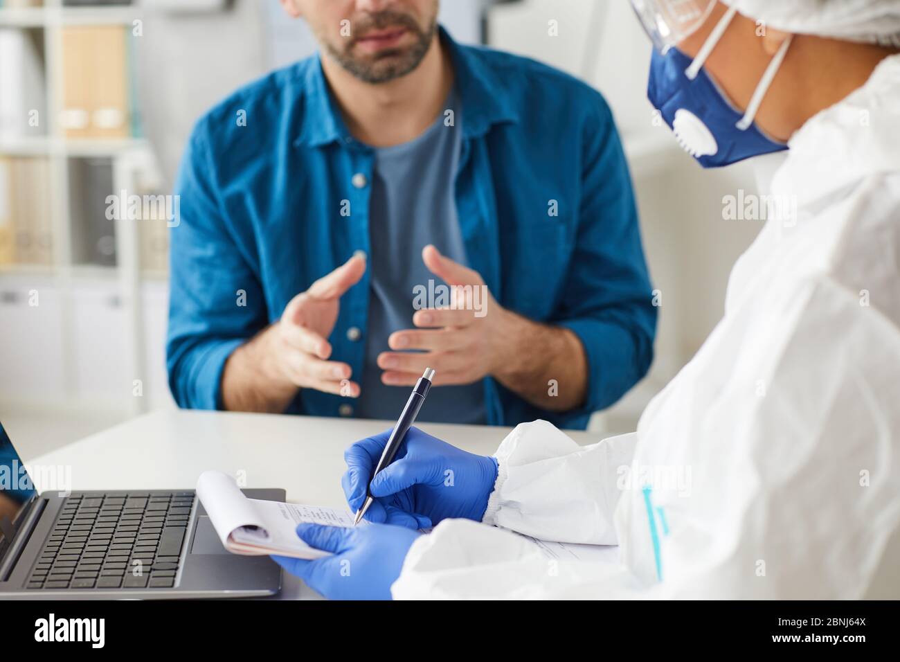 Primo piano del medico in indumenti protettivi seduto al tavolo e la prescrizione di scrittura con il paziente in background Foto Stock