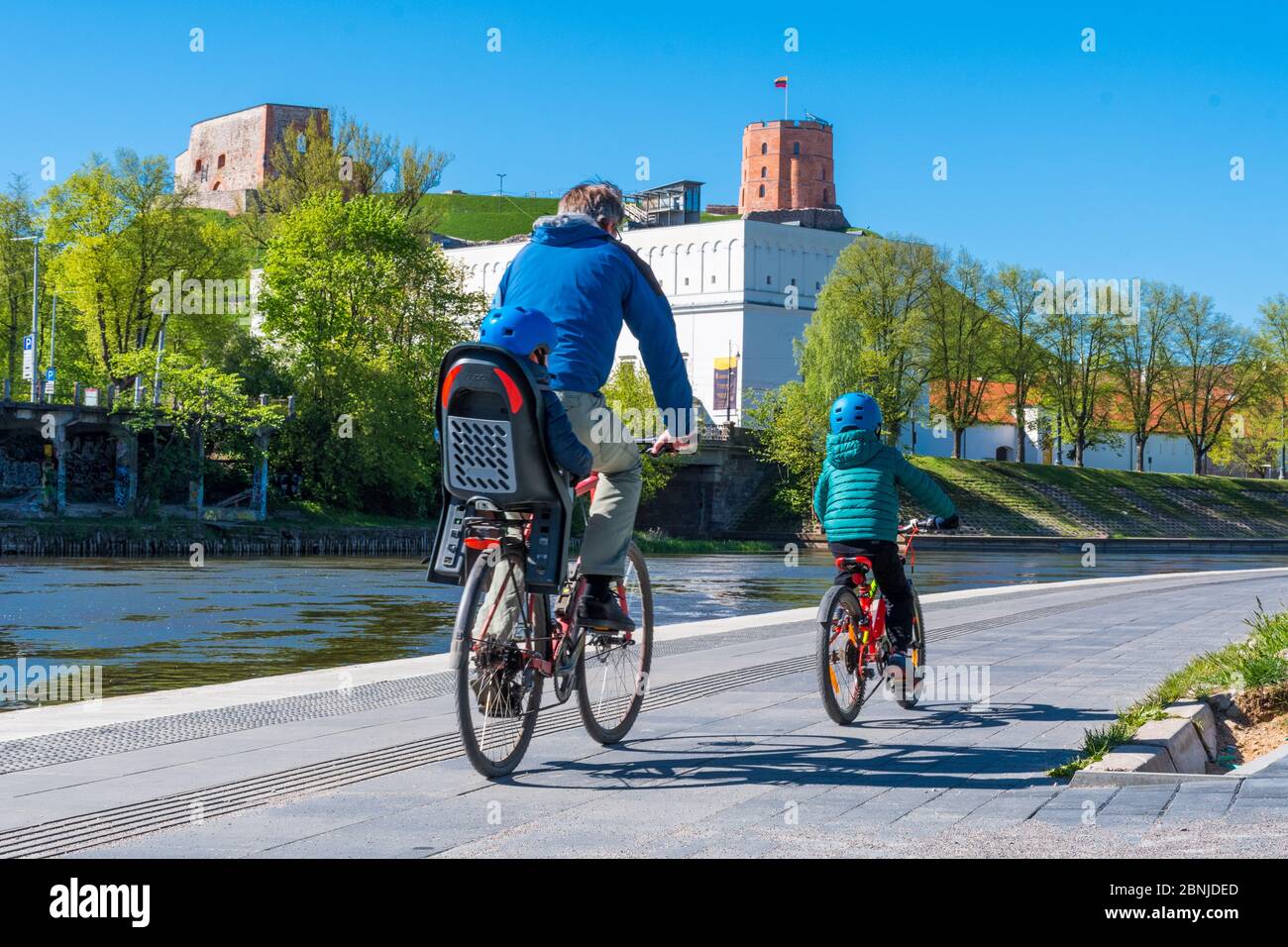 Padre e bambini in bicicletta su strada con parco, fiume e castello sullo sfondo durante l'emergenza Covid o Coronavirus, trasporto sostenibile Foto Stock