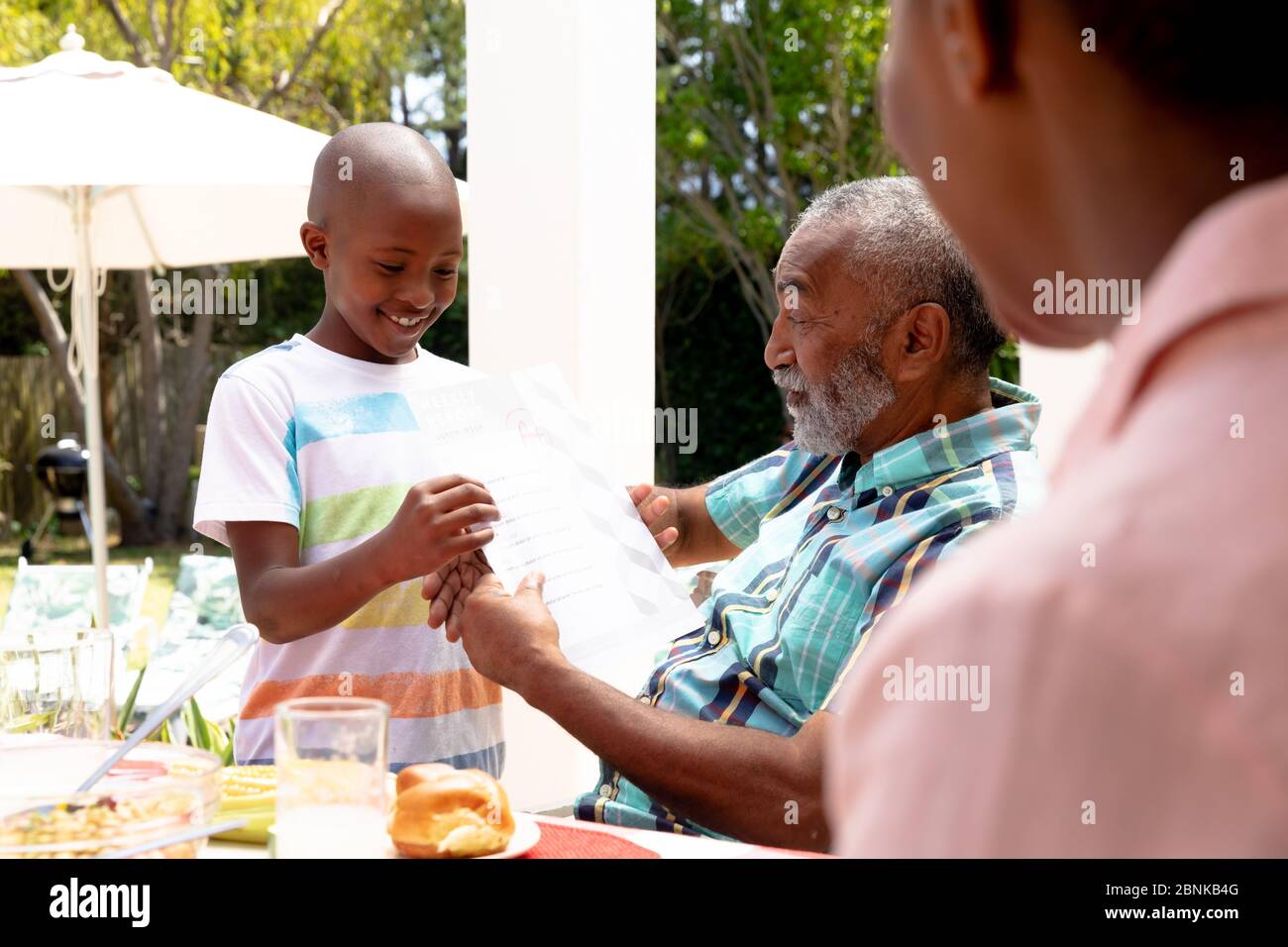 Ragazzo afroamericano che mostra al nonno i suoi compiti durante un pranzo di famiglia in giardino Foto Stock