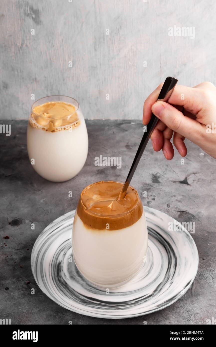 Bevanda popolare caffè coreano Dalgon. Il cucchiaio lungo mescola la schiuma montata di caffè solubile, zucchero e acqua calda con il latte in vetro su sfondo grigio Foto Stock