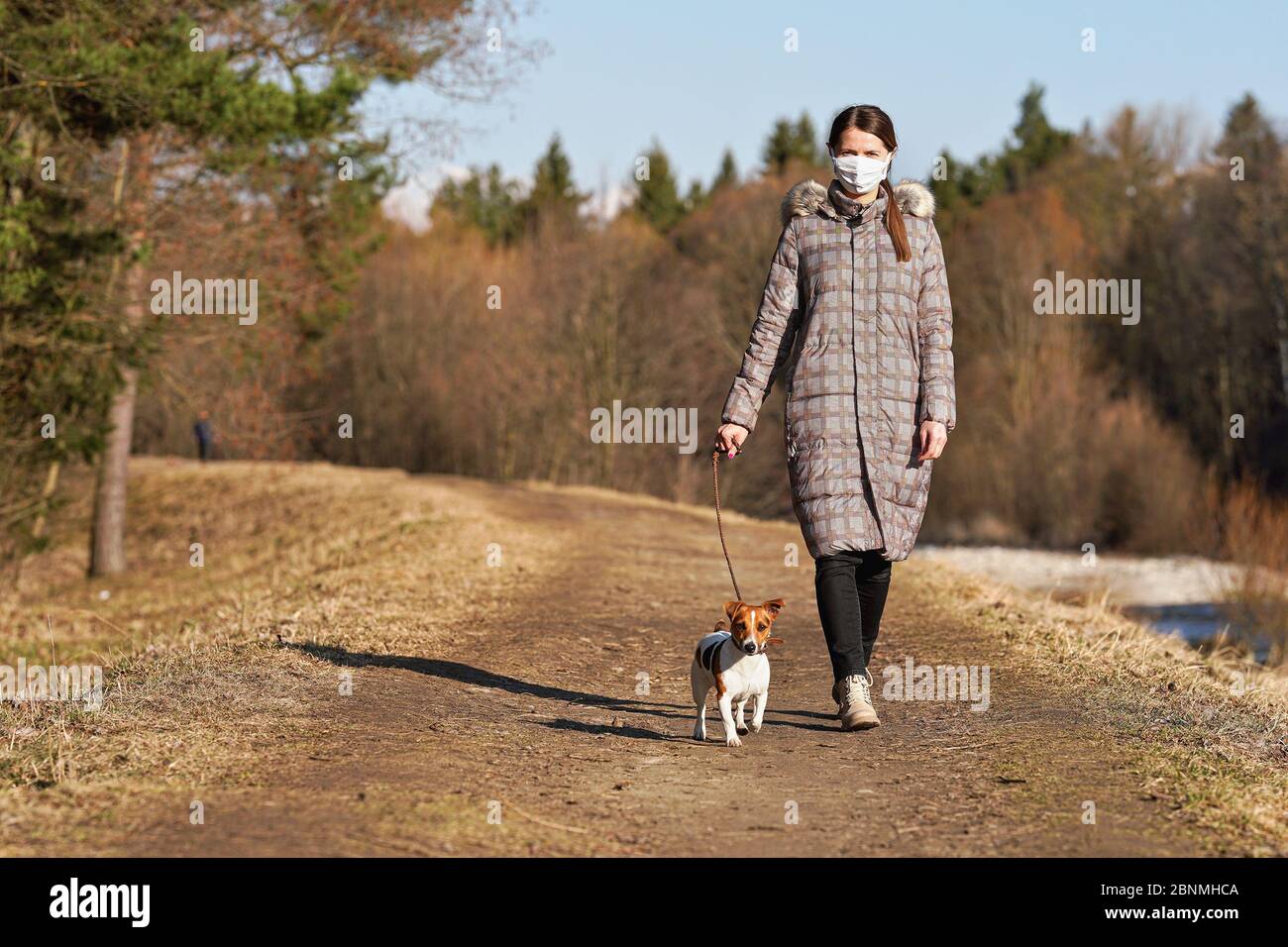 Giovane donna in giacca calda, con la maschera virus viso bocca, cammina il suo cane su strada di campagna. Le maschere sono obbligatorie fuori casa durante il covid di coronavirus- Foto Stock