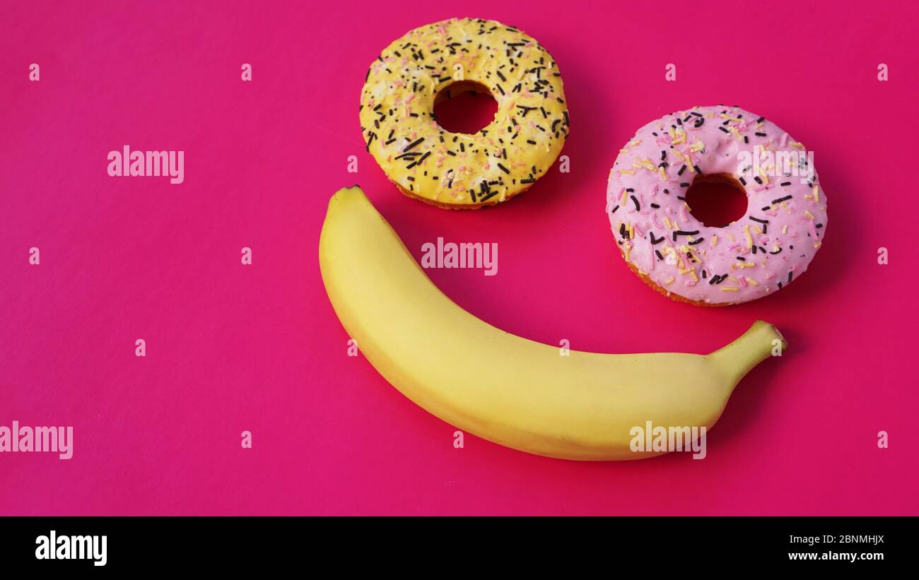 Due ciambelle e una banana giacciono sulla superficie rosa, formando un'astrazione dell'emoticon - sorriso. Preso dal punto più alto Foto Stock