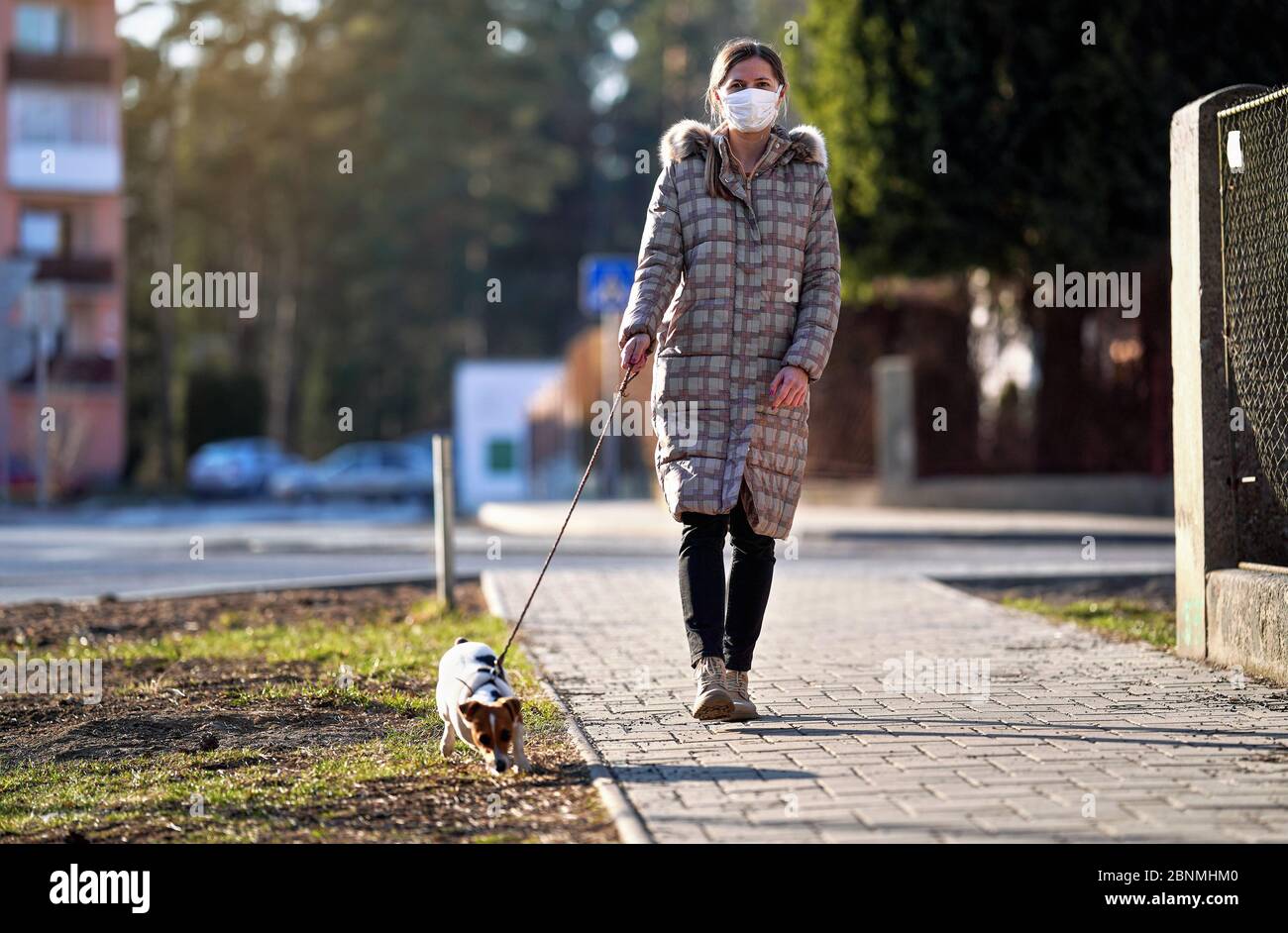 Giovane donna in giacca calda, con la maschera virus bocca faccia, cammina il suo cane sul marciapiede. Le maschere sono obbligatorie fuori casa durante il coronavirus covid-19 out Foto Stock