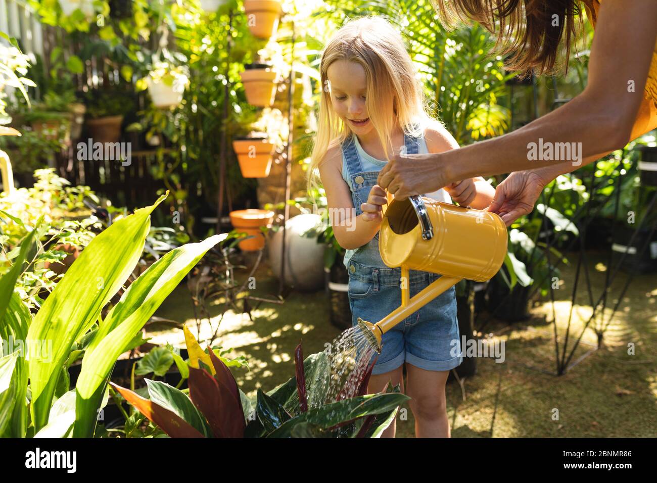 Una donna caucasica e sua figlia annaffiatura piante con annaffiatoio in un giardino soleggiato Foto Stock