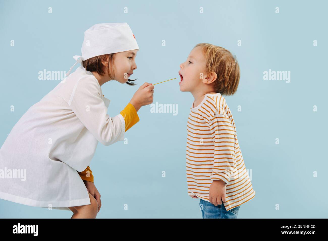 Bambina che gioca un medico, controllando la gola del ragazzo su sfondo blu. Foto Stock