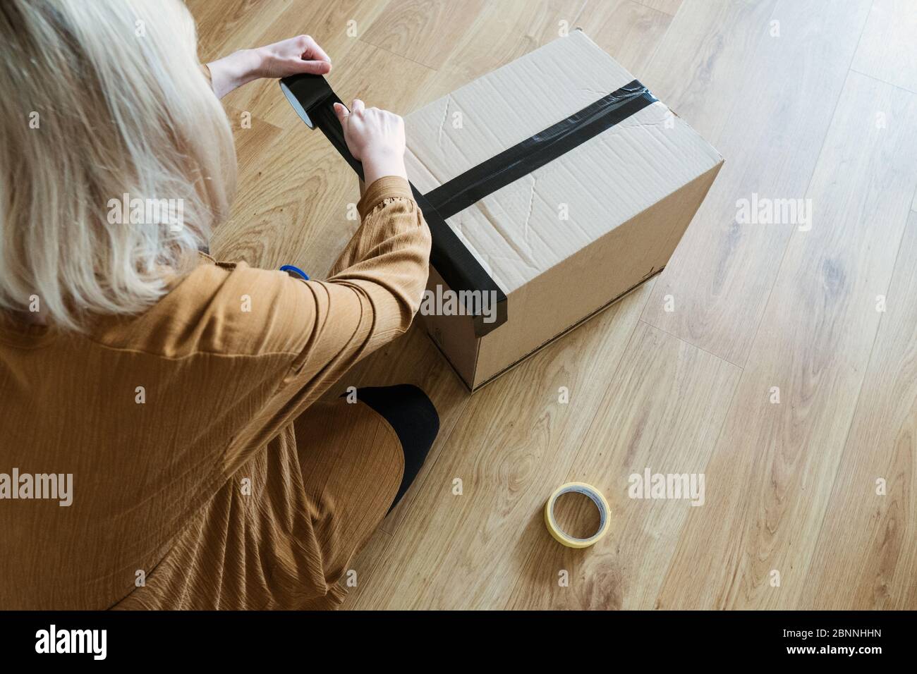 Una giovane donna che è un cliente prepara un pacchetto per la spedizione utilizzando nastro e forbici. Consumatore ragazza che tiene scatola di cartone sul pavimento. Foto Stock