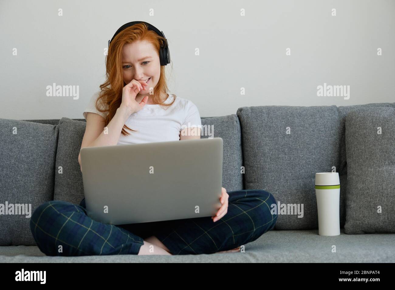 Ragazza rossa in abiti e cuffie a casa si siede sul divano, gambe incrociate e sorridenti, guardando il computer portatile Foto Stock