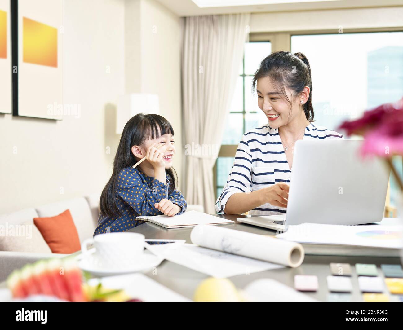 giovane designer asiatica madre che lavora da casa utilizzando un computer portatile mentre si prende cura di figlia (grafica in background alterato digitalmente) Foto Stock