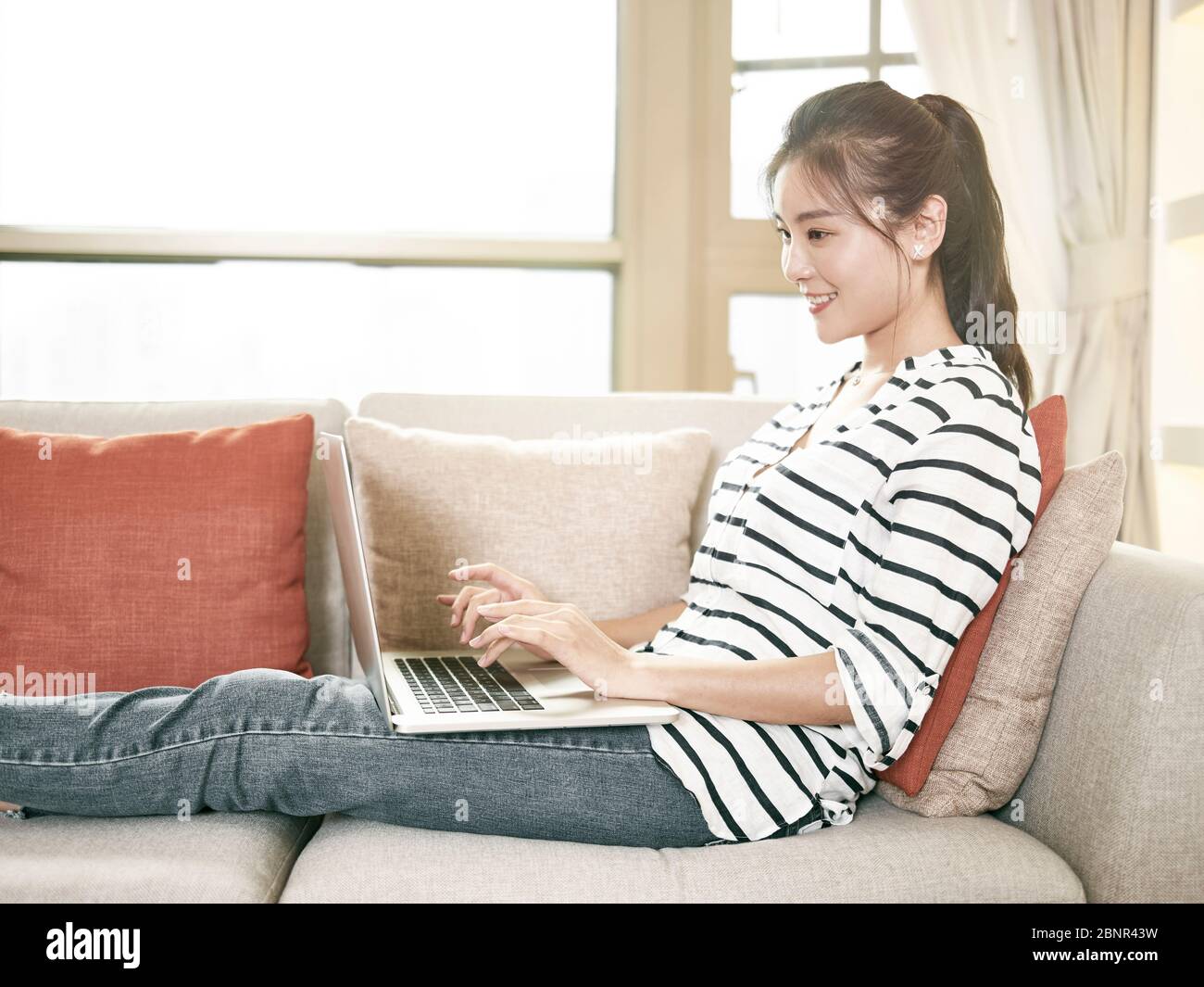 giovane donna asiatica che lavora da casa seduta sul divano usando un computer portatile Foto Stock
