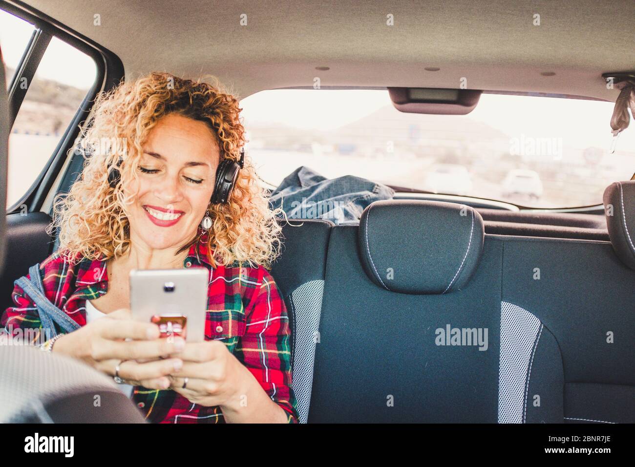 Allegra donna felice sedersi sui sedili posteriori all'interno di una moderna auto utilizzando un telefono cellulare per ascoltare musica con le cuffie - persone che si godono viaggi con concetto di tecnologia Foto Stock