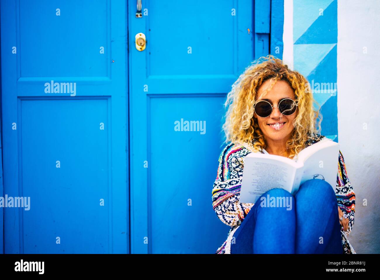 Ritratto di immagine blu di beuaitufl felice adulta donna lettura e godendo di un libro sedersi sul pavimento con casa e porta in background - persone che studiano e leggono in attività di svago all'aperto Foto Stock