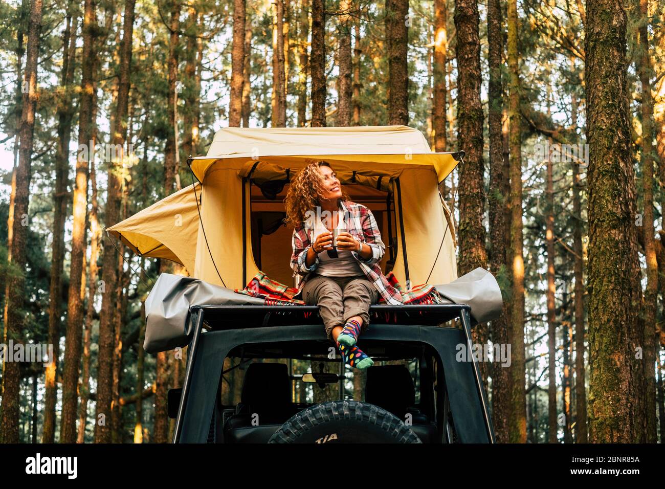 Viaggio e wanderlust stile di vita concetto con felice donna adulta solitaria sedersi sul tetto tenda auto veicolo con legno foresta in background godendo la natura e vacanze all'aperto Foto Stock