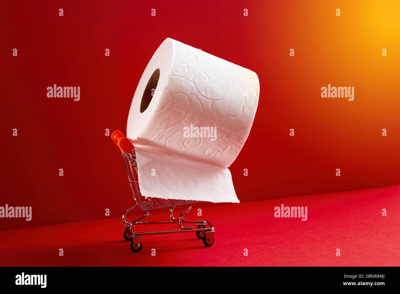 Rotolo di carta igienica bianca con un carrello della spesa in miniatura su sfondo rosso. Concetto di acquisto antipanico. Foto Stock