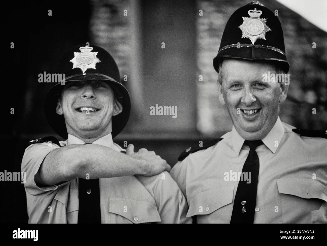 Due costabili di polizia maschile che si propongono per una risata con caschi custoditi di dimensioni sbagliate. Inghilterra, Regno Unito. Circa anni 80 Foto Stock