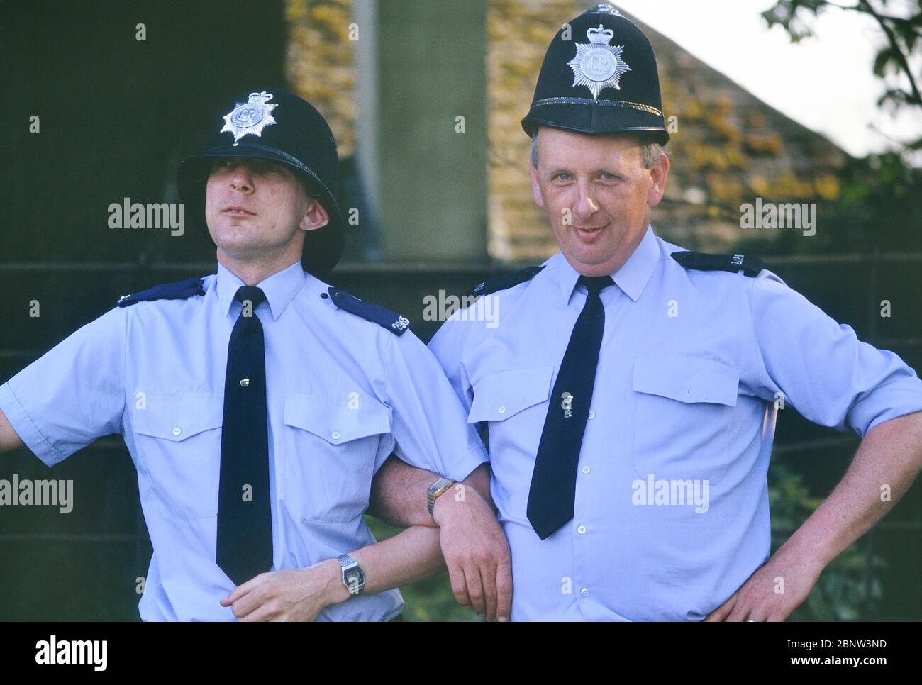 Due costabili di polizia maschile che si propongono per una risata con caschi custoditi di dimensioni sbagliate. Inghilterra, Regno Unito. Circa anni 80 Foto Stock