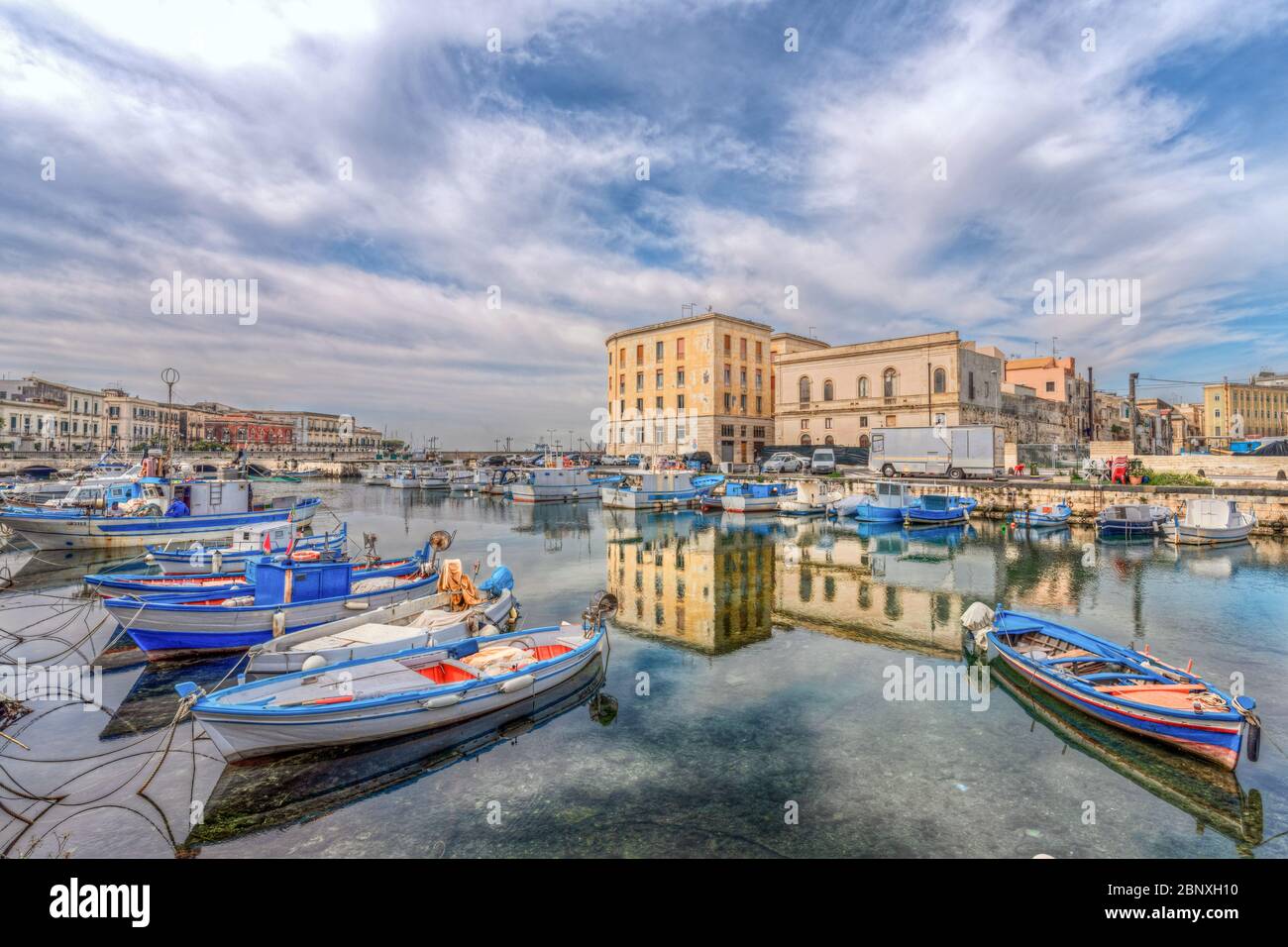 Siracusa Sicilia/Italia - Aprile 11 2020: Le barche dei pescatori ormeggiate nel canale del molo di Ortigia e il palazzo si riflettono nelle sue acque Foto Stock