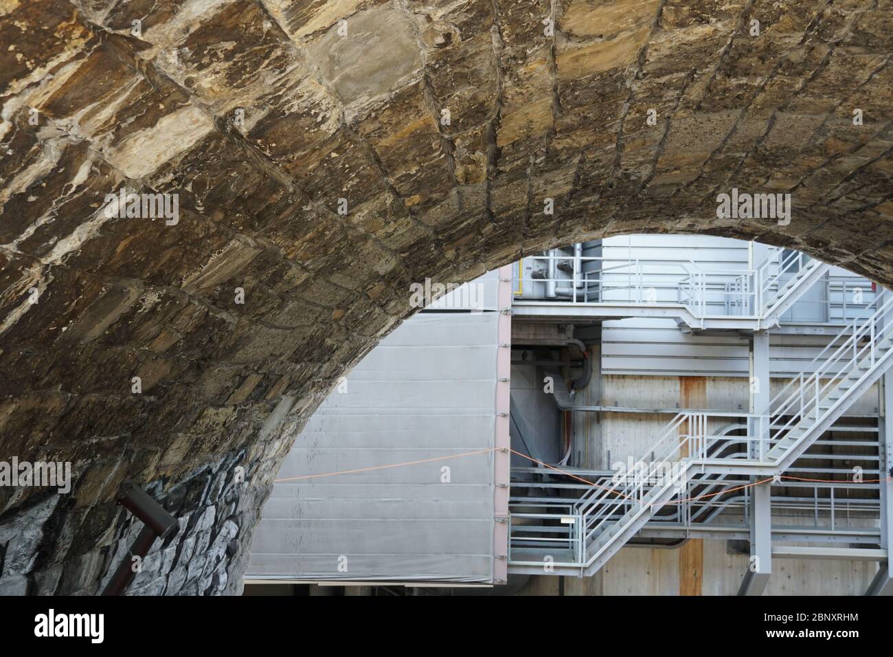 arco in cubetti di pietra che dà vista ad un tratto posteriore di un edificio industriale con tubi e scala metallica Foto Stock