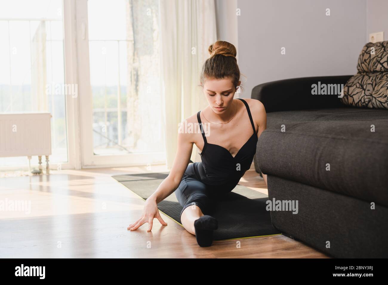 Giovane donna sportiva siede sul tappetino in spago diviso, ballerina professionista, allenatore, indossando la lebbrosa nera, attività a casa durante l'isolamento Foto Stock