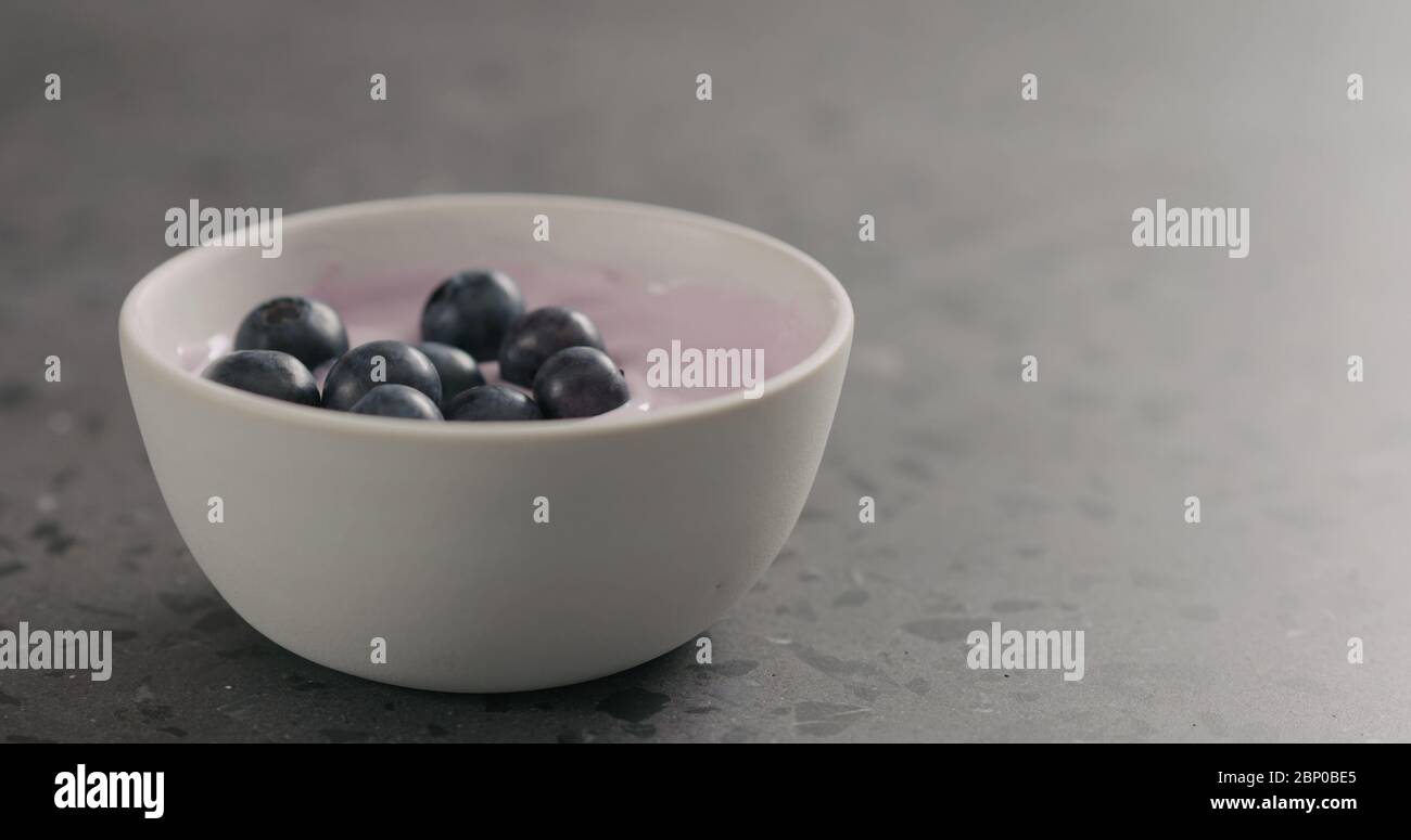 i mirtilli maturi cadono nello yogurt al mirtillo in una ciotola bianca con spazio di copia Foto Stock