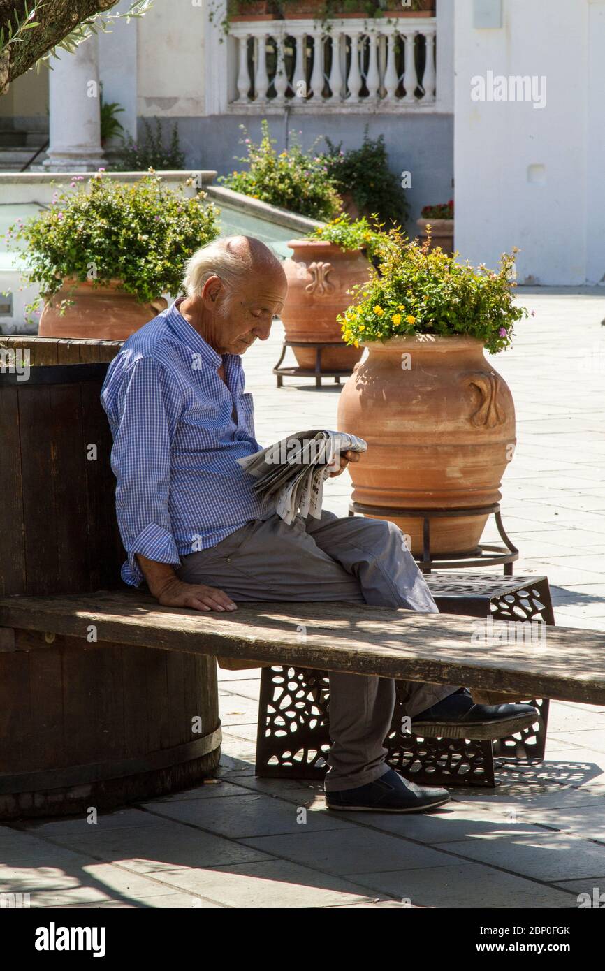 Uomo solo seduto su una panchina di cortile, all'ombra di un albero, leggendo un giornale Foto Stock