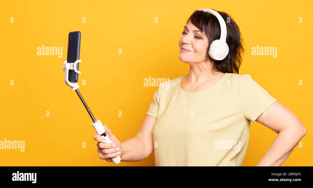 Bella donna senior che effettua videochiamate con smartphone, monopod e cuffie wireless su sfondo giallo. Foto Stock