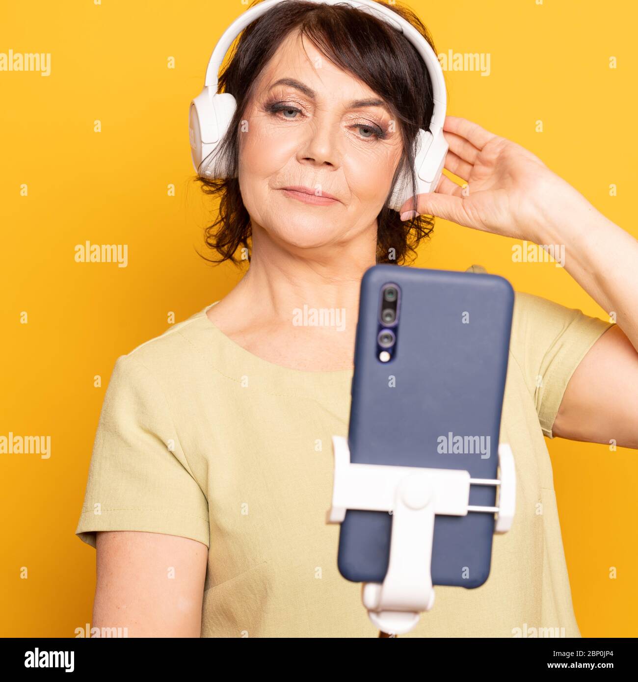 Bella donna senior che effettua videochiamate con smartphone, monopod e cuffie wireless su sfondo giallo Foto Stock
