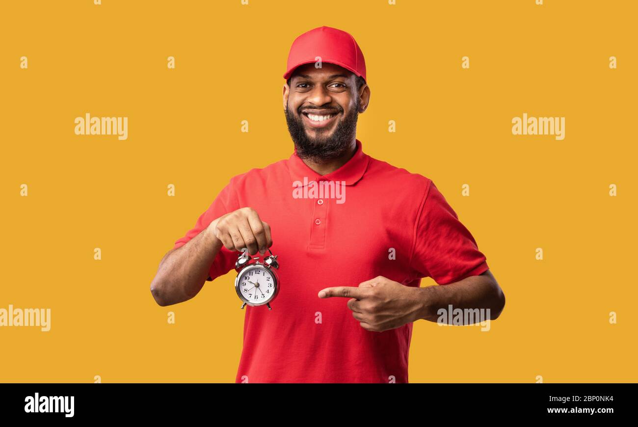 Corriere che punta la barretta all'orologio sorridendo alla telecamera, sfondo giallo Foto Stock
