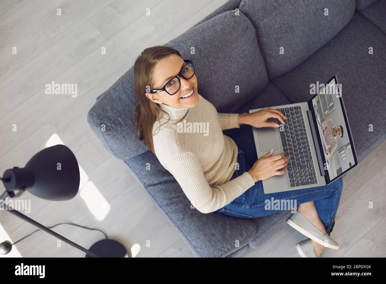 Lavoro online a casa. Una donna sorridente con un computer portatile guarda la videocamera in una videochiamata con un partner seduto su un divano in un soggiorno. Foto Stock