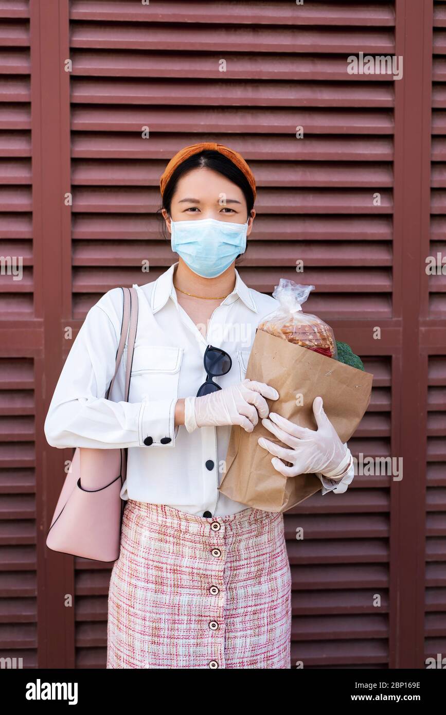 Donna asiatica che tiene una borsa della spesa con generi alimentari mentre si indossa maschera e guanti per prevenire la diffusione del virus durante la pandemia di coronavirus con copy space ag Foto Stock