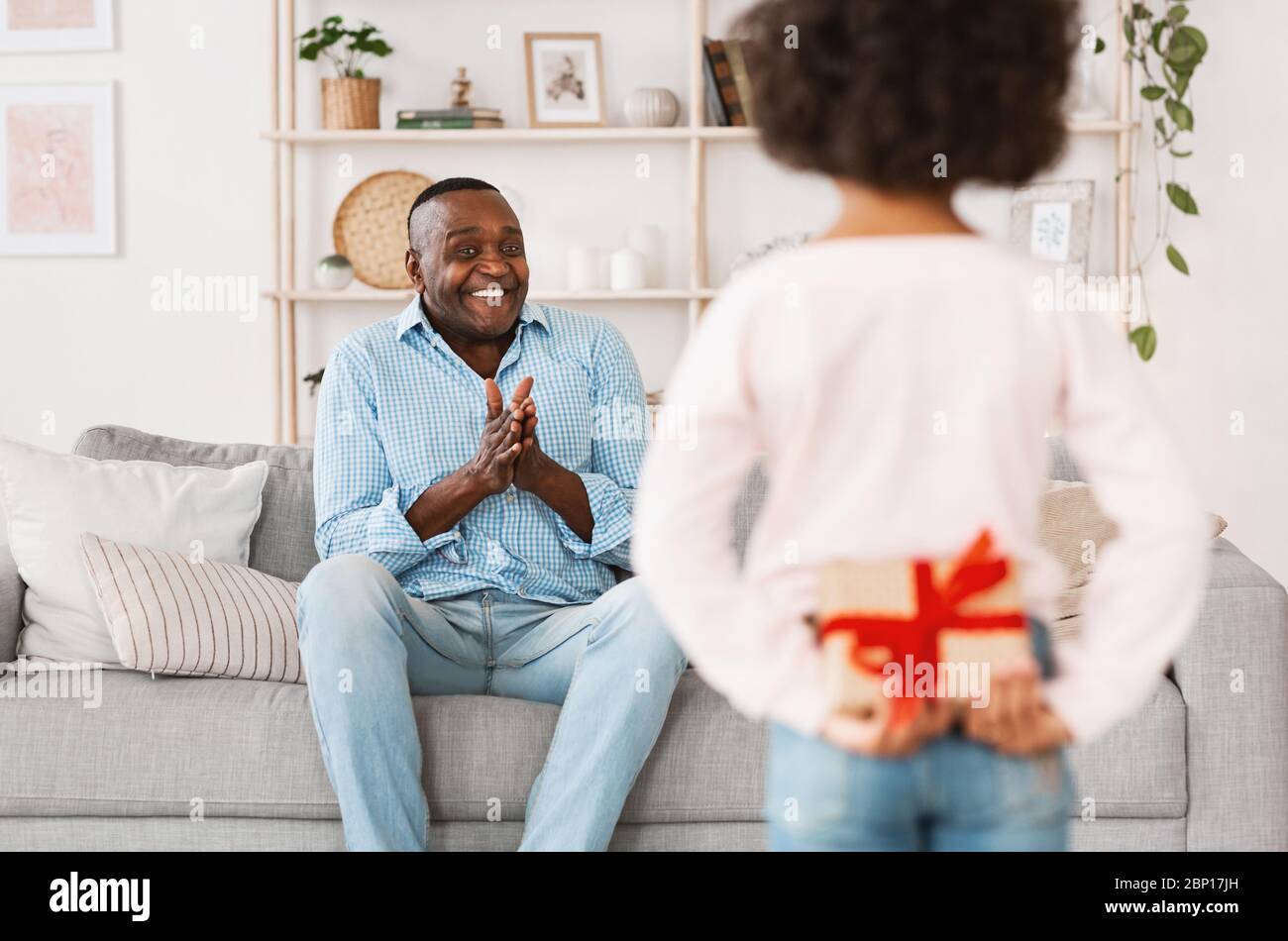 Rimani a casa e festeggia. Piccolo bambino afro-americano che tiene presente per nonno dietro la schiena in interni Foto Stock