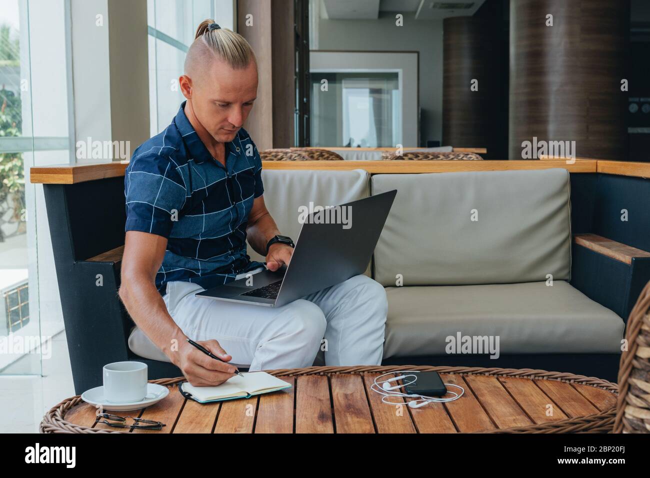 Un giovane uomo d'affari di successo in una camicia e pantaloni è in un bar, occupato con il lavoro remoto utilizzando la tecnologia moderna. Funziona con il computer portatile, controllando il report Foto Stock