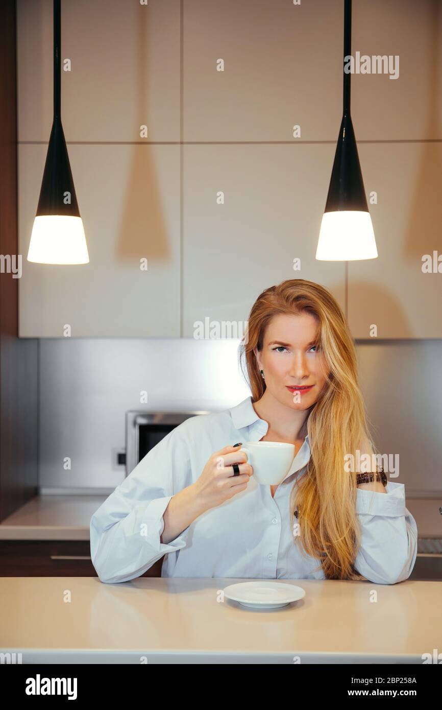 Bella femmina con capelli biondi lunghi tiene una tazza di tè/caffè in mano mentre ci guarda. Sta in piedi ad un tavolo o ad un bar, possibilmente in una cucina. Foto Stock