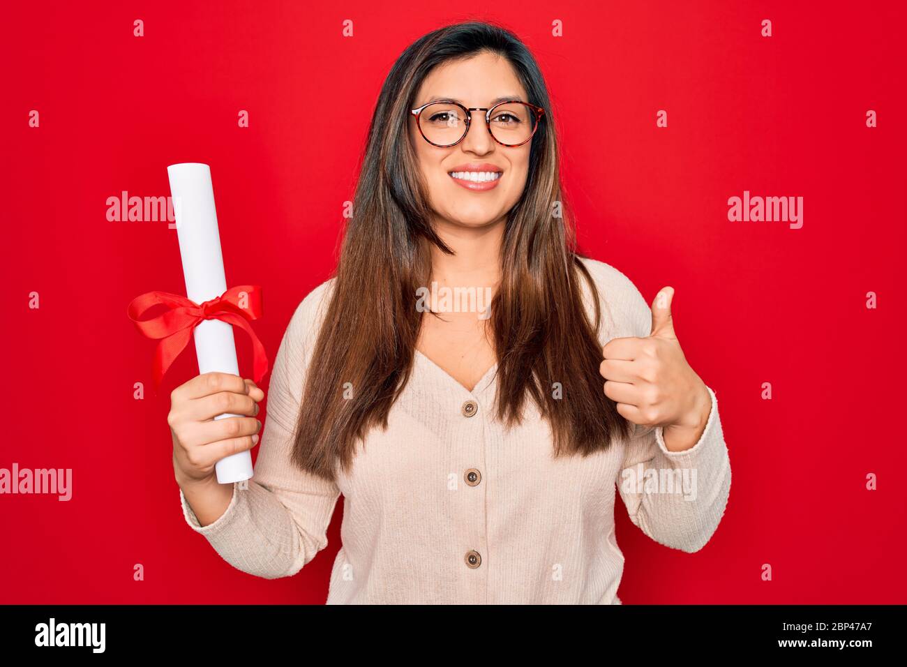 Giovane donna intelligente ispanica indossare occhiali tenendo laurea università su sfondo rosso felice con grande sorriso facendo ok segno, pollice su con le dita, ex Foto Stock